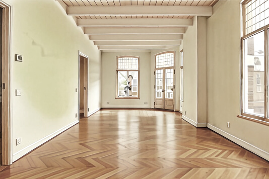 Das Bild zeigt einen hellen, geräumigen Raum mit Holzfußboden und großen Fenstern.